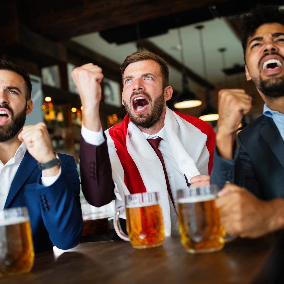 Men fans screaming and watching football on TV and drink beer in a pub